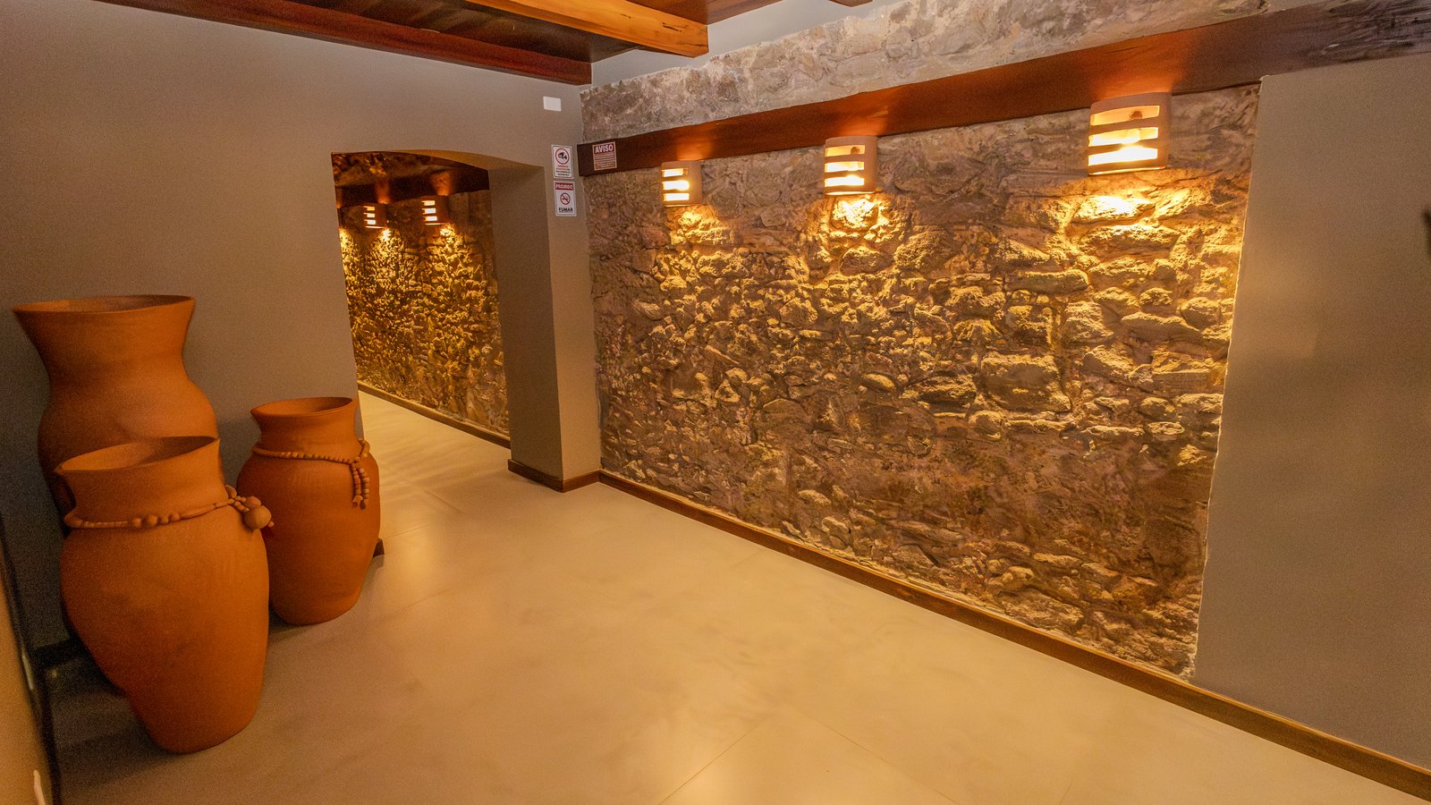 Corredor interno com parede de pedra iluminada e vasos de barro